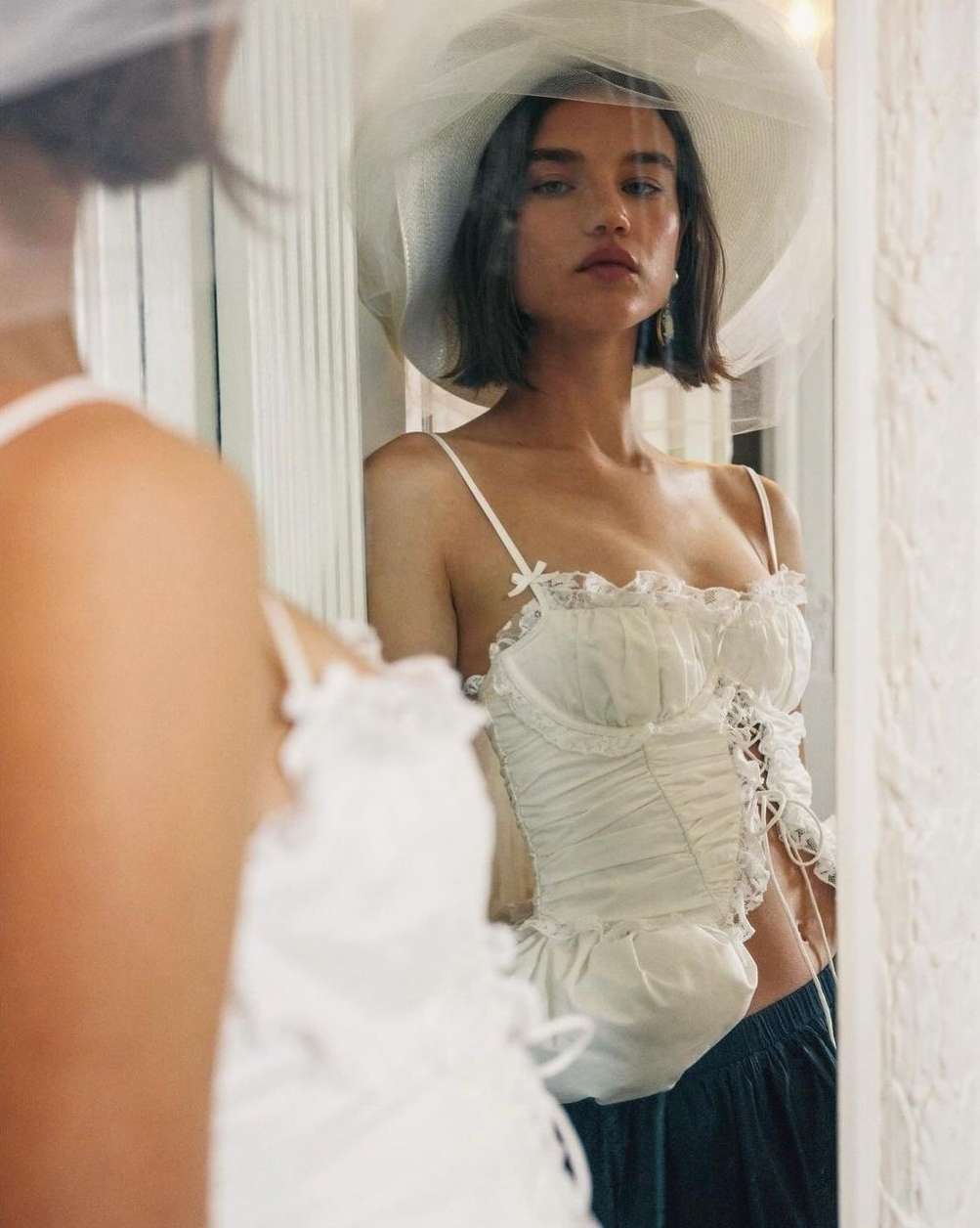 Young adult woman with short hair and a hat with netting, posing in a lacy corset top, reflected in a mirror, indoor setting.