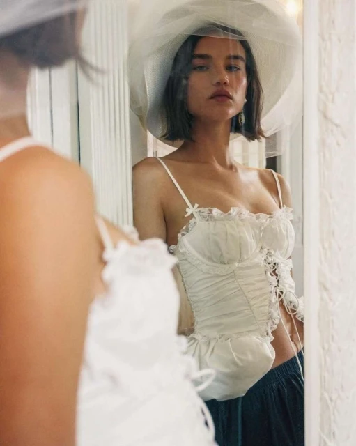 Young adult woman with short hair and a hat with netting, posing in a lacy corset top, reflected in a mirror, indoor setting.