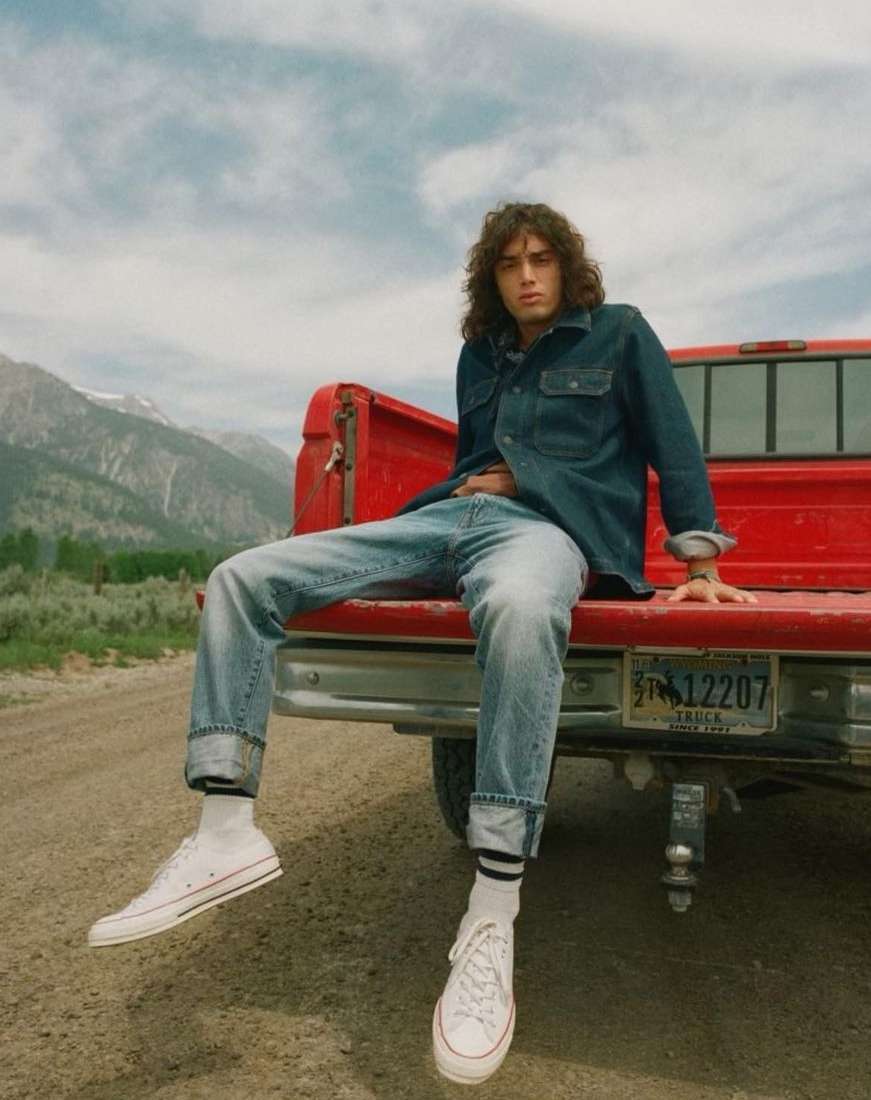 Young man in denim outfit sits on the back of a red pickup truck with a mountain landscape and blue sky in the background.
