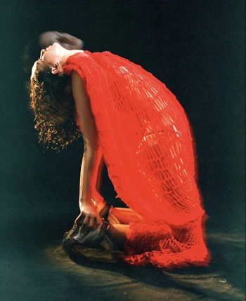 A woman in a bright red textured dress kneeling and arching her back against a dark background, photographed by Bryan Rodner Carr