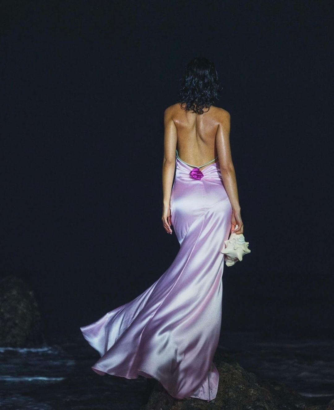 Woman in lilac gown holds seashell on a rocky beach. She faces the ocean, wearing a backless dress under dark, moody lighting.