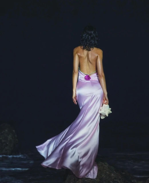 Woman in lilac gown holds seashell on a rocky beach. She faces the ocean, wearing a backless dress under dark, moody lighting.