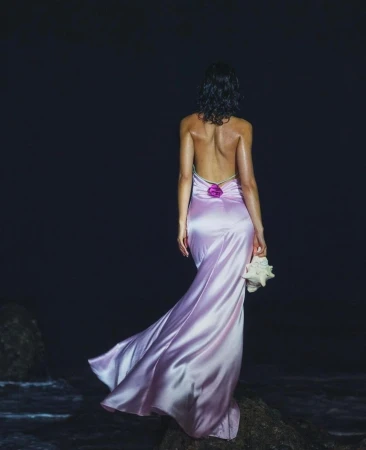 Woman in lilac gown holds seashell on a rocky beach. She faces the ocean, wearing a backless dress under dark, moody lighting.
