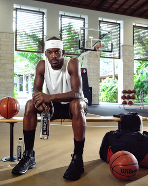 A male basketball player sits on a bench on an indoor court with basketballs, a sports bag, and Essentia water bottles.