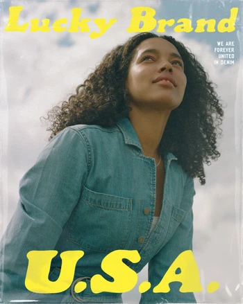 Portrait of a woman with curly hair wearing a denim jumpsuit against a cloudy sky with yellow typography