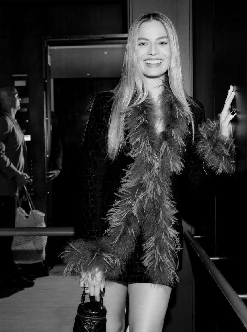Blonde woman in a black dress with a feathered boa and a small purse, posing indoors in a dimly lit environment with other people.