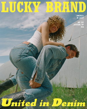 A couple in denim share a playful moment against a rural backdrop. The woman sits on the man's shoulders in a lighthearted outdoor fashion shot.