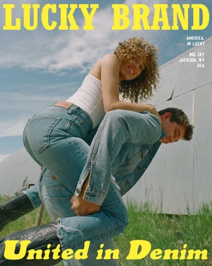 A couple in denim share a playful moment against a rural backdrop. The woman sits on the man's shoulders in a lighthearted outdoor fashion shot.