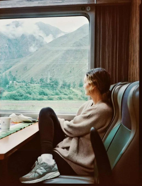 Woman in sweater looks out train window at Peruvian mountain landscape. She sits at a table with a coffee and wears New Balance sneakers.