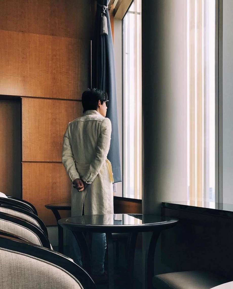 Adult male stands looking out window in hotel room interior. Light wood walls, drapery, modern chair and table, light from window.