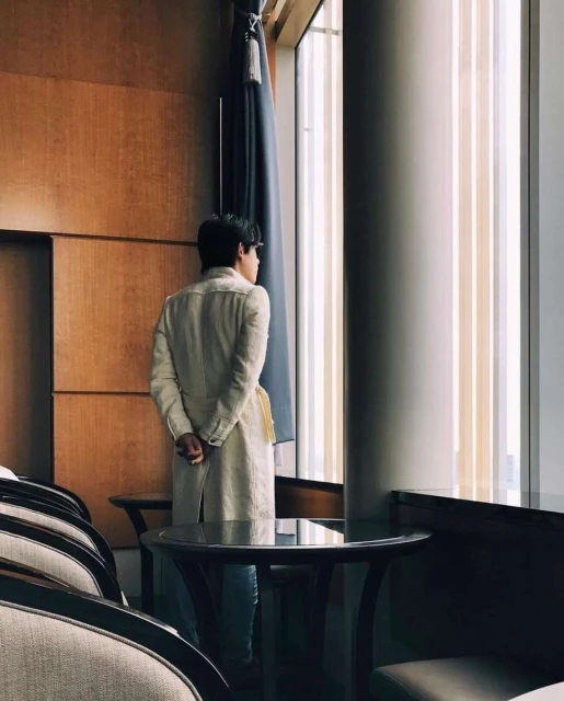 Adult male stands looking out window in hotel room interior. Light wood walls, drapery, modern chair and table, light from window.
