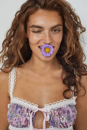 A woman winks with a purple flower in her mouth, wearing a floral tank top against a white backdrop in an indoor setting.