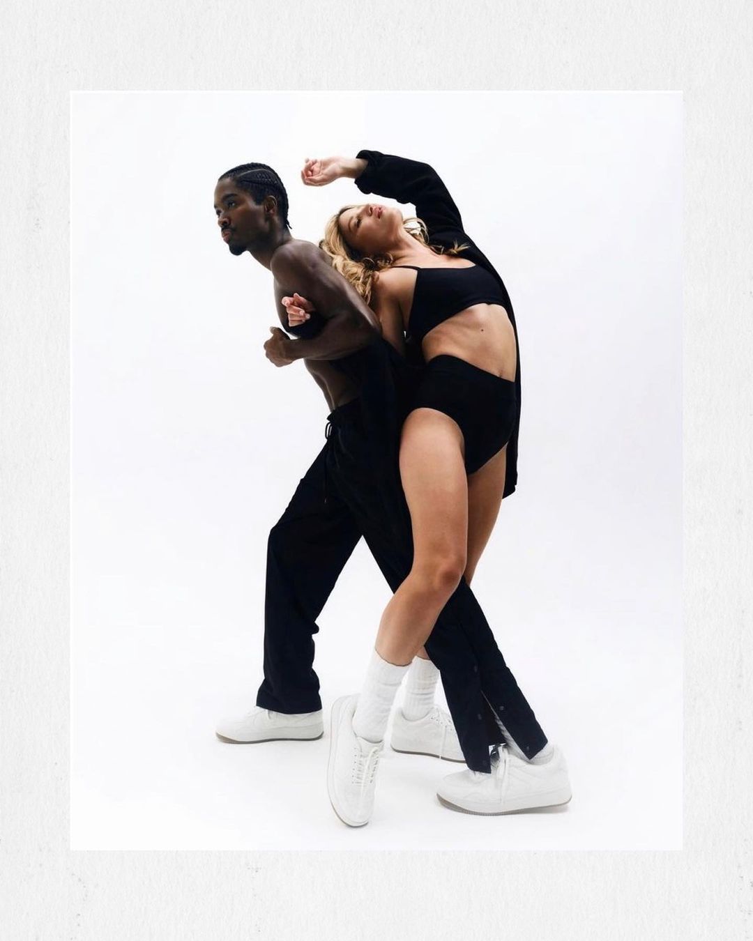 A man and woman in black athletic clothing pose on a white background in a studio setting, showcasing a modern, minimalist fashion style.