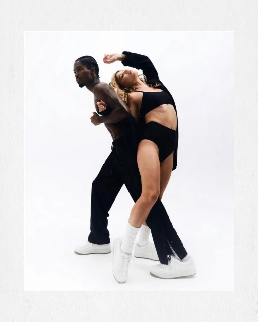 A man and woman in black athletic clothing pose on a white background in a studio setting, showcasing a modern, minimalist fashion style.