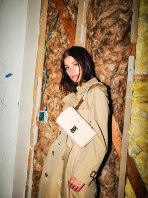 A young woman poses indoors wearing a trench coat and carrying a handbag with a logo, against an unfinished wall backdrop.