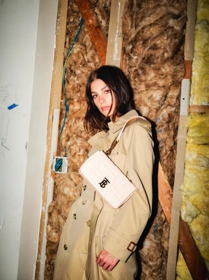 A young woman poses indoors wearing a trench coat and carrying a handbag with a logo, against an unfinished wall backdrop.