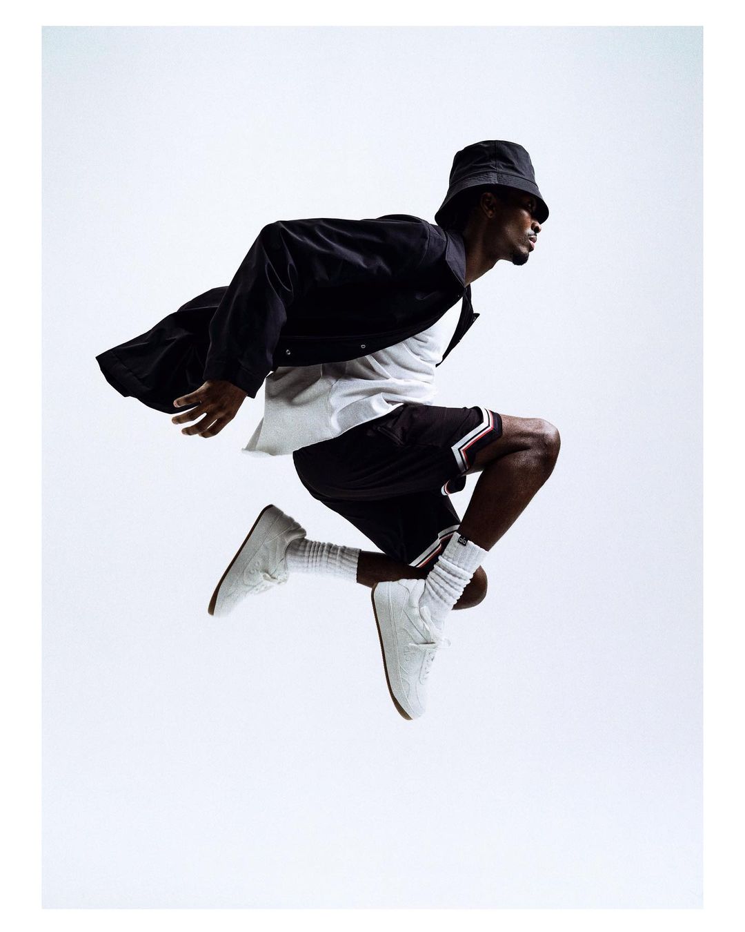 Full body shot of an adult male in mid-air wearing casual clothes, a bucket hat, jacket, shorts, socks, and sneakers, against a white background.