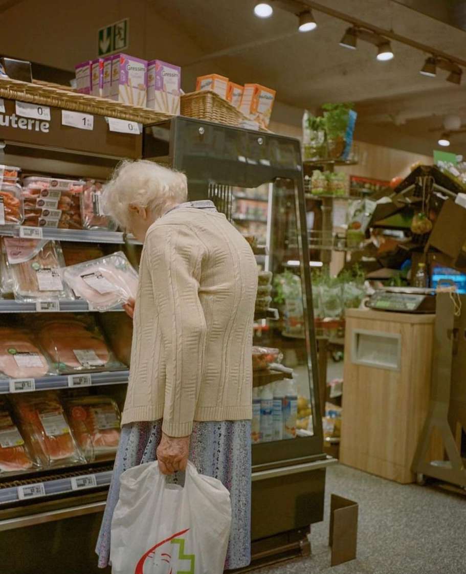 Senior woman shops for packaged meat at a grocery store, wearing a cardigan and holding a shopping bag in an indoor setting.