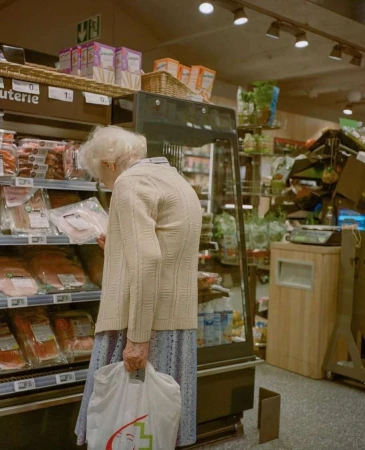 Senior woman shops for packaged meat at a grocery store, wearing a cardigan and holding a shopping bag in an indoor setting.