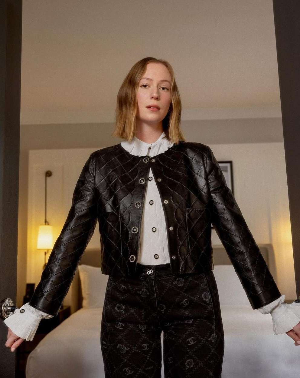 A stylish adult woman with light-brown hair is standing indoors wearing a black leather jacket, patterned pants, and a white collared shirt.