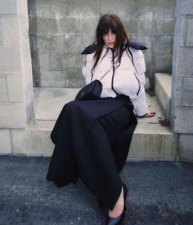 Woman in white puffer jacket and black skirt seated outdoors against a concrete block wall by Bryan Rodner Carr