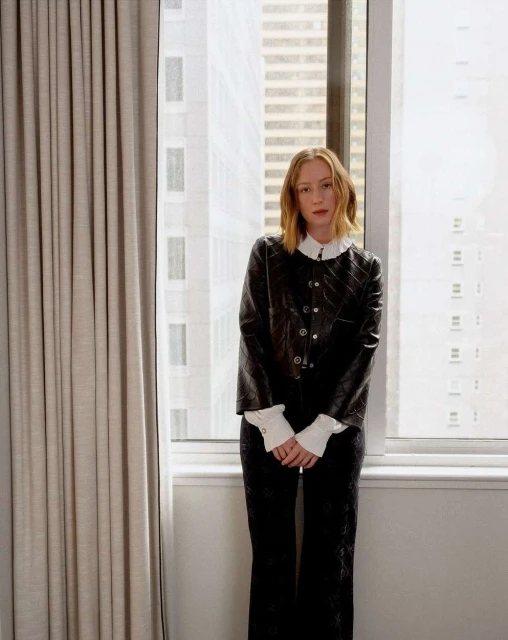 A slender woman with short blonde hair is posing in a black jacket and pants against a backdrop of neutral curtains and a city view.