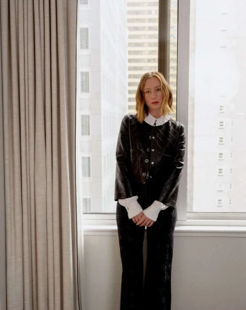 A slender woman with short blonde hair is posing in a black jacket and pants against a backdrop of neutral curtains and a city view.