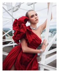 Cara Delevingne in a red one-shoulder dress with a large shoulder ruffle posing on a white metal rooftop structure.
