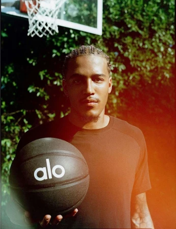 A young man holding a black basketball with alo branding outdoors in front of a basketball hoop and greenery.