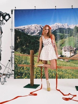 Model standing before an alpine backdrop banner wearing a white patterned dress and long red ribbons.