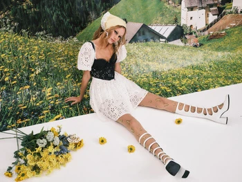 Model in white dress with black bodice and yellow headscarf seated on white platform before alpine meadow backdrop