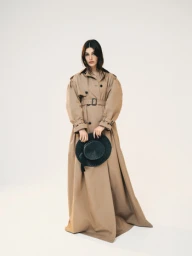 Model in a long beige belted trench coat holding a dark hat against a plain white studio background.