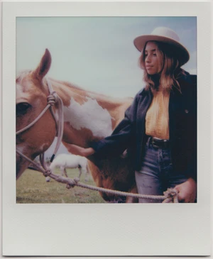 Polaroid photograph of a woman wearing a wide-brim hat standing next to a horse in a field.