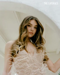 Portrait of a young woman with long wavy brown hair wearing a feather-embellished dress framed by a white arched architecture element.