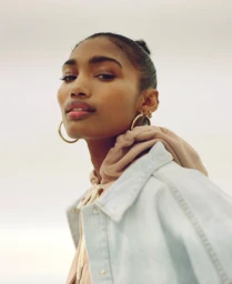 Portrait of a woman wearing a light blue denim jacket and hoop earrings against a neutral sky background