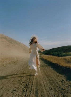 Woman in a white dress running along a dusty dirt road flanked by hills under a clear blue sky