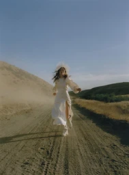 Woman in a white dress running along a dusty dirt road flanked by hills under a clear blue sky