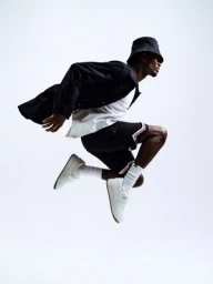 Black man mid-air jumping wearing a black bucket hat, dark jacket, white t-shirt, shorts, and white sneakers against a white background.