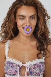 Portrait of a woman with curly brown hair holding a purple flower in her mouth against a white background