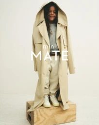 Young child wearing an oversized beige trench coat standing on a wooden crate against a white background.
