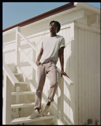 Black man wearing a white t shirt and beige pants leaning on white wooden stairs against a white wall.