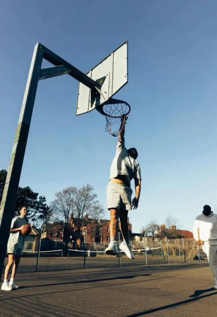 Action shot of a man dunking a basketball on an outdoor court under a clear sky, with two other people standing nearby.