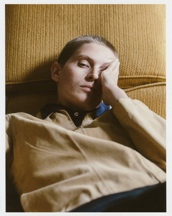 Eye-level portrait of a light-skinned adult with short hair reclining against a textured yellow couch, looking at the camera with a hand on their face.