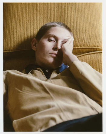 Eye-level portrait of a light-skinned adult with short hair reclining against a textured yellow couch, looking at the camera with a hand on their face.