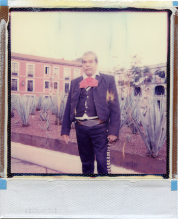 Adult man in a mariachi suit stands in a courtyard with agave plants and a pink building in the background in a vintage-style photo.