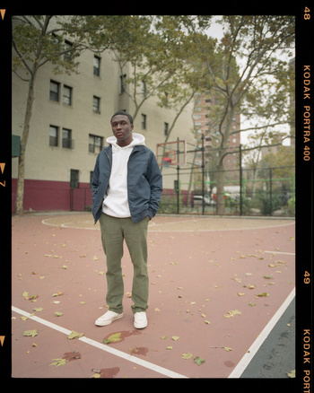 A young man stands in the center of a weathered outdoor basketball court. He is dressed in a casual jacket, hoodie, and trousers.