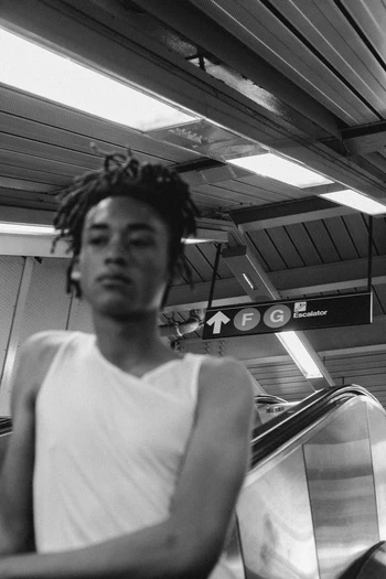Young adult with dreadlocks in a white tank top stands near an escalator with illuminated signs in an urban subway station, captured in black and white.