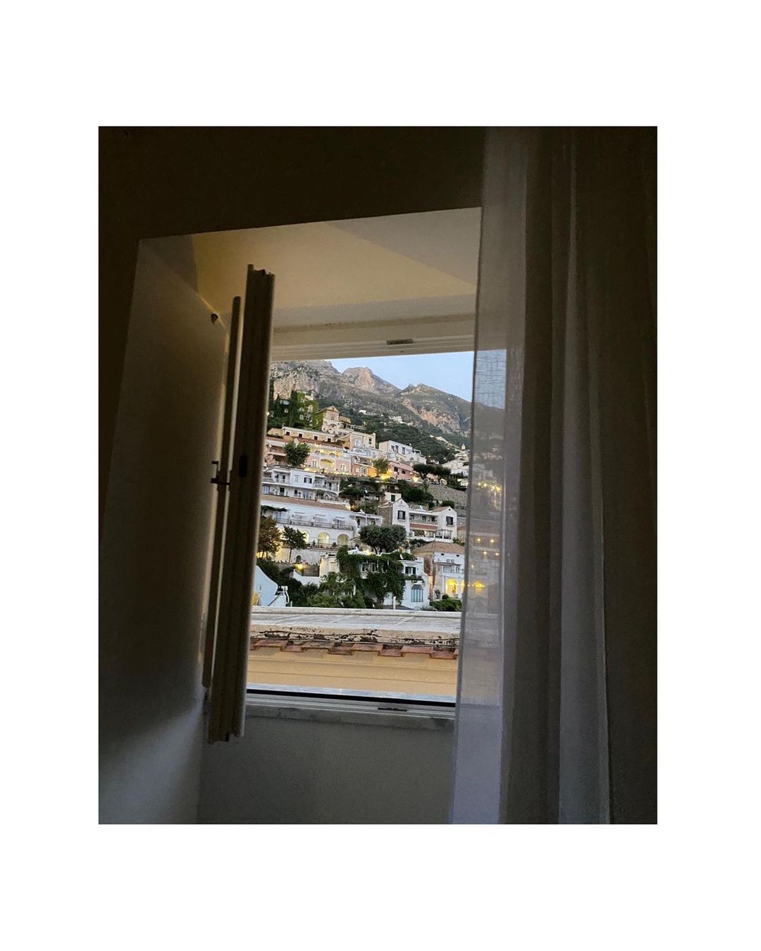 Scenic view through open window, featuring hillside buildings in warm light with mountain backdrop, partially obscured by sheer white curtain.