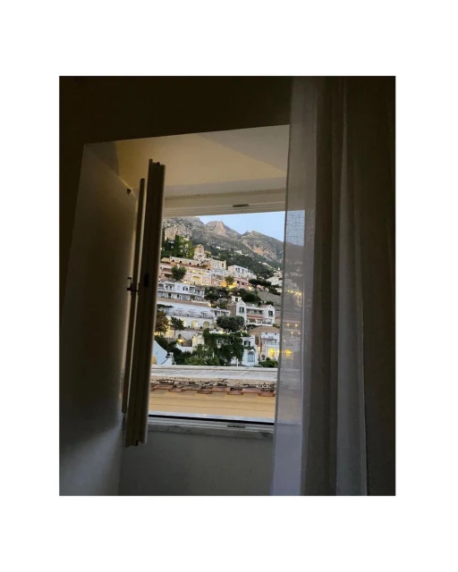Scenic view through open window, featuring hillside buildings in warm light with mountain backdrop, partially obscured by sheer white curtain.