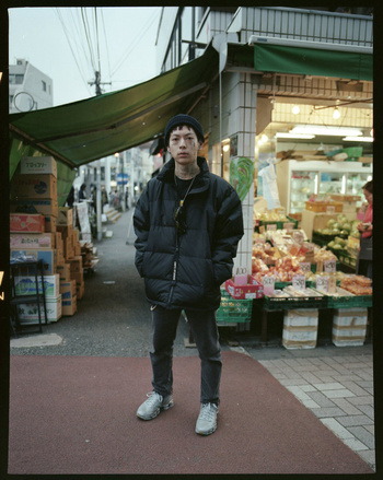 Portrait of a person with tattoos and a beanie in a black puffer jacket standing outside of a market stall on a city street.